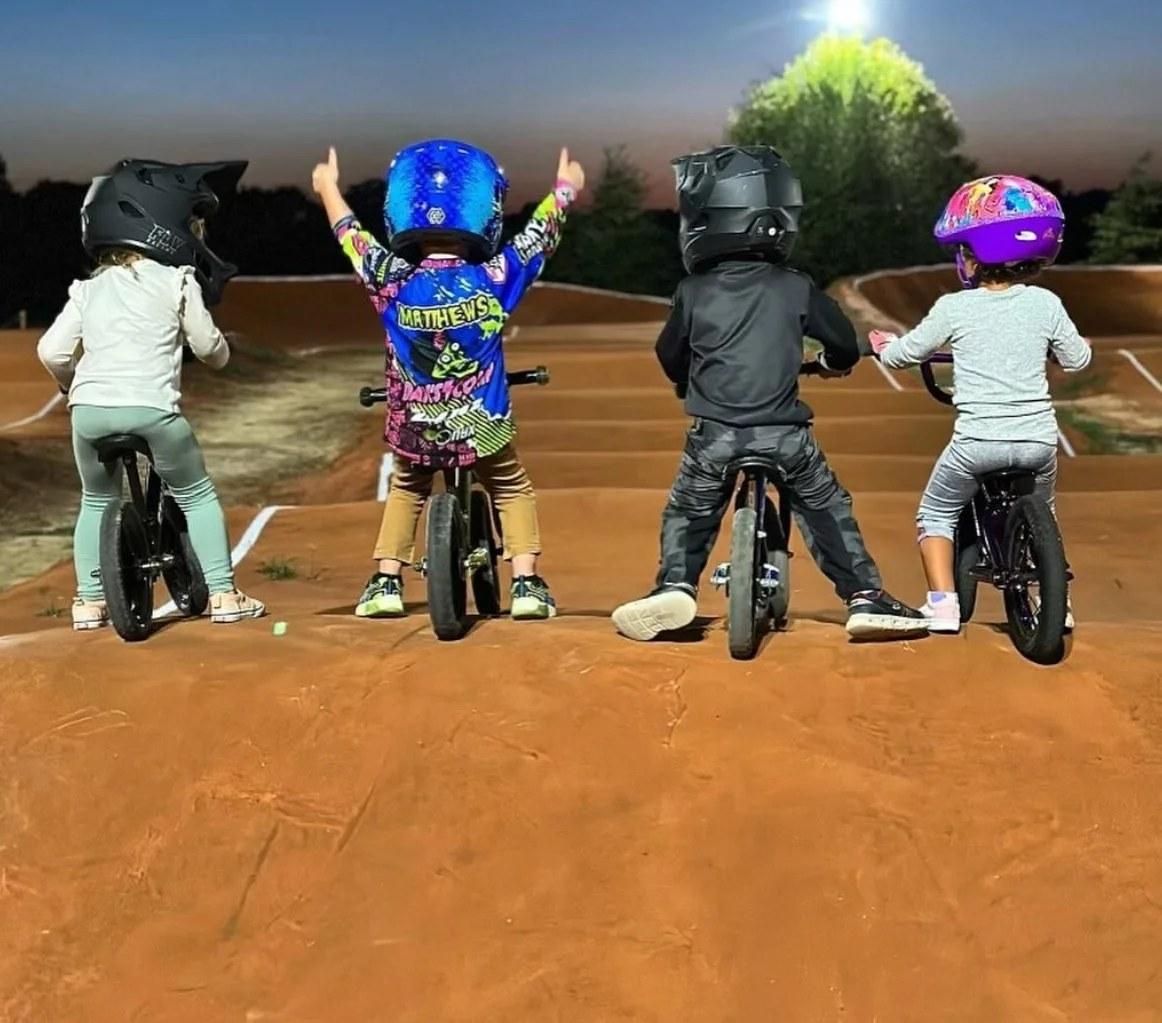 Four balance bike riders at the top of the start hill at dusk, one with both arms raised