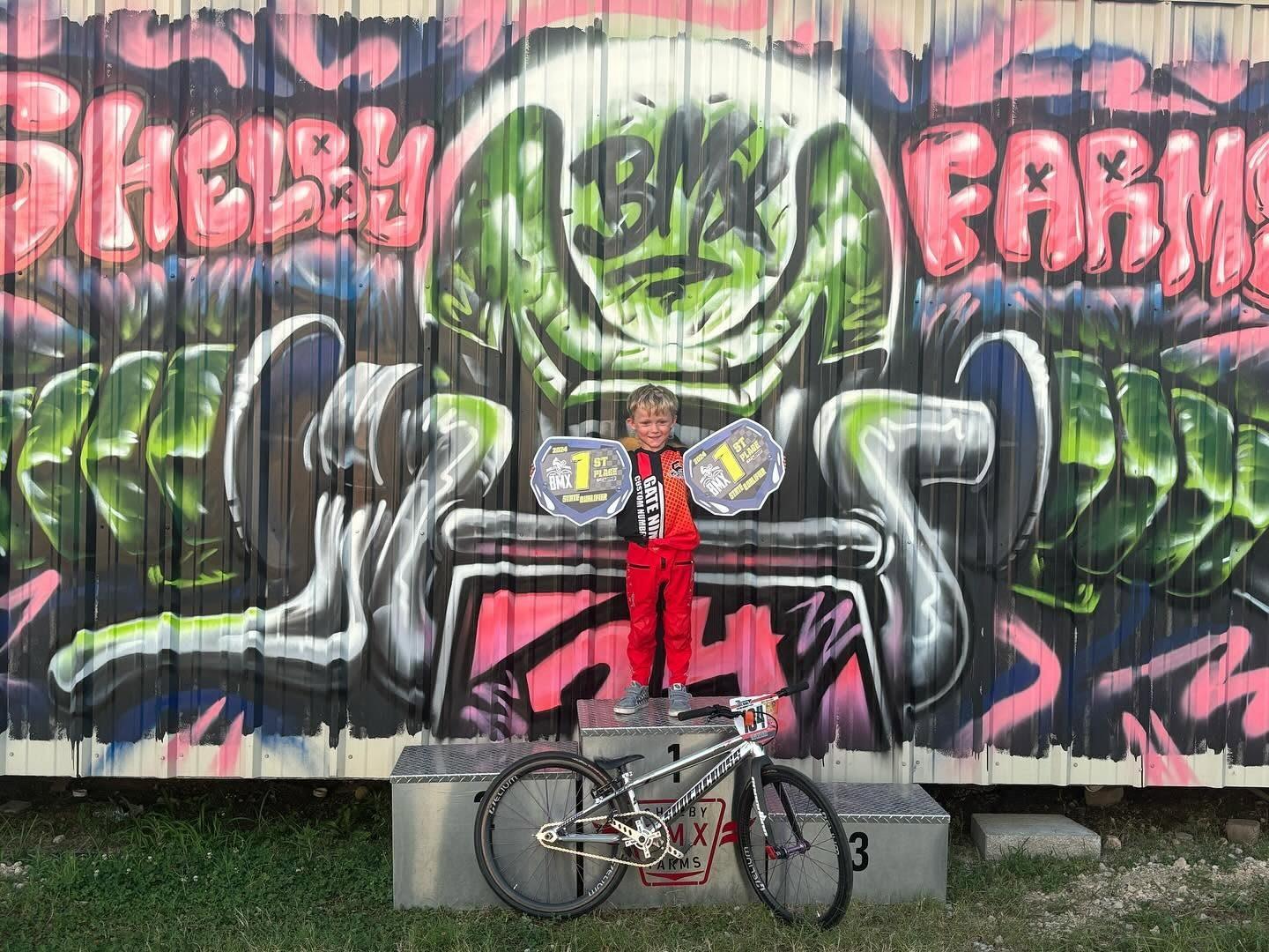 A young rider on the podium in front of the Shelby Farms BMX mural, holding two first place plates